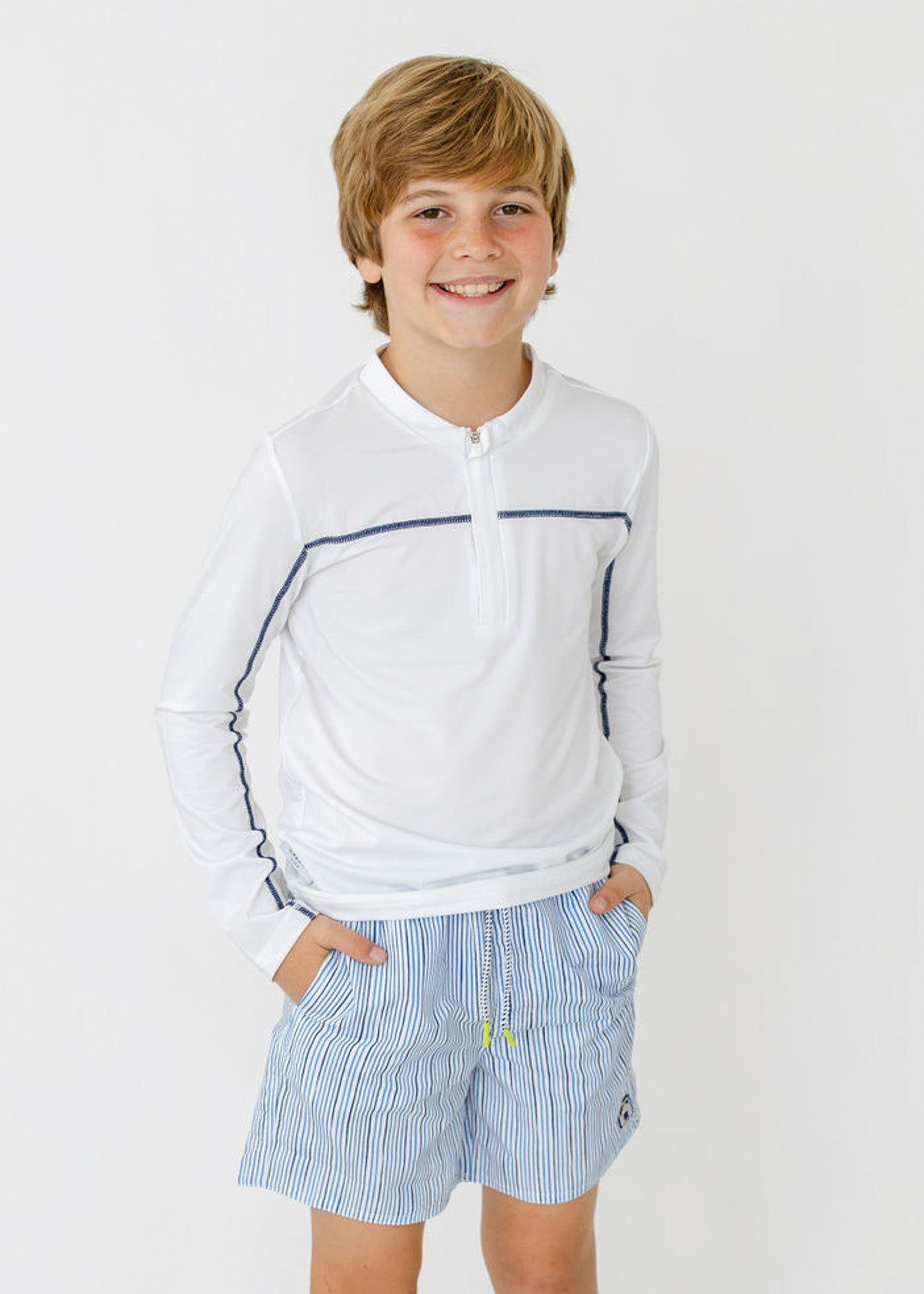 A boy wearing the sun protective Boys White Sport Zip Top and Boys Seaside Stripe Swim Trunk from Cabana Life.