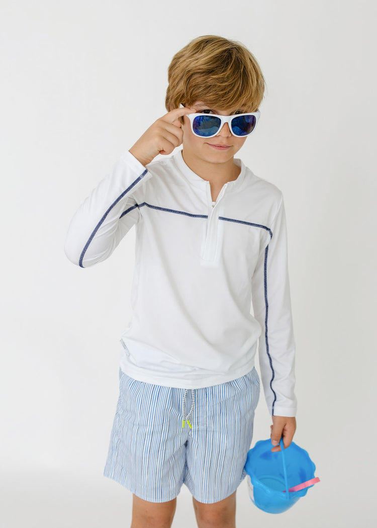 A boy holding a beach pail and shovel while wearing sunglasses and the sun protective Boys White Sport Zip Top and Boys Seaside Stripe Swim Trunk from Cabana Life.