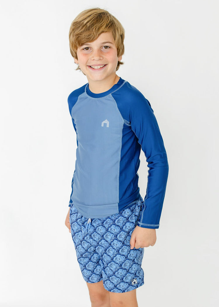 A boy wearing the sun protective Boys Blue Colorblock Long Sleeve Rashguard with hands in pockets of the Boys Seaside Swim Trunks from Cabana Life.