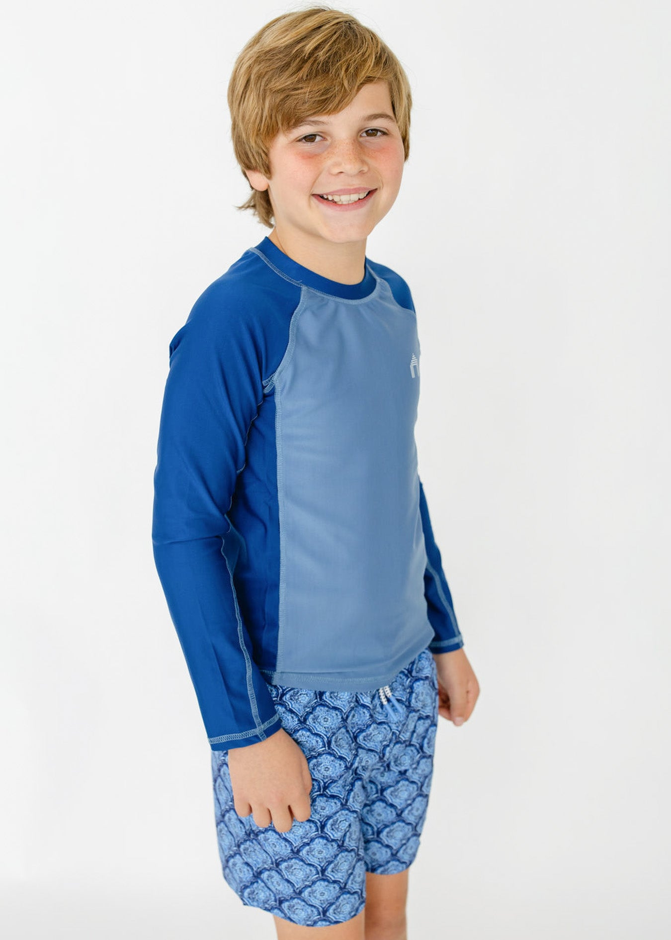 A boy wearing the sun protective Boys Blue Colorblock Long Sleeve Rashguard and Boys Seaside Swim Trunks from Cabana Life.