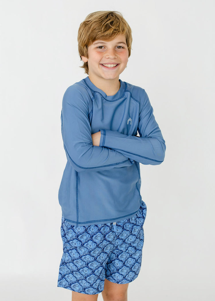 A blonde boy wearing the sun protective Boys Light Blue Long Sleeve Rashguard and Boys Seaside Swim Trunks from Cabana Life.