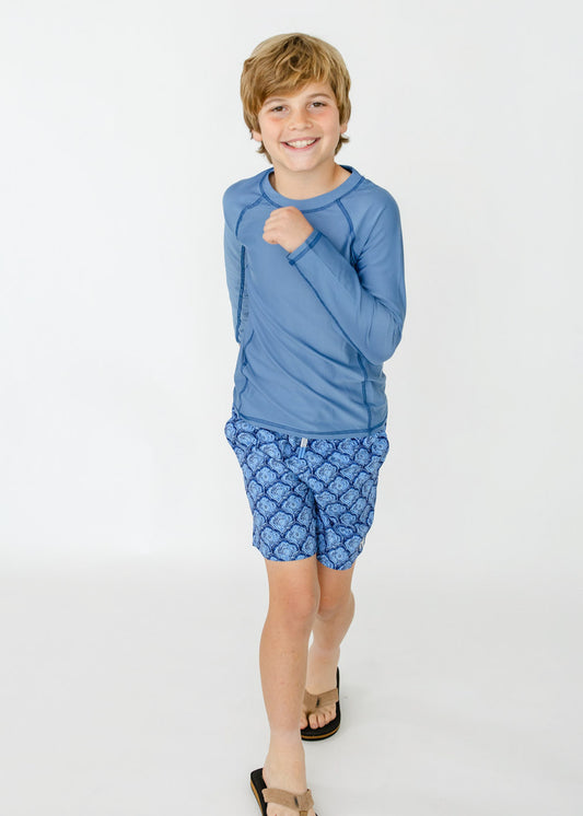 A blonde boy running in flip flops wearing the sun protective Boys Light Blue Long Sleeve Rashguard and Boys Seaside Swim Trunks from Cabana Life.