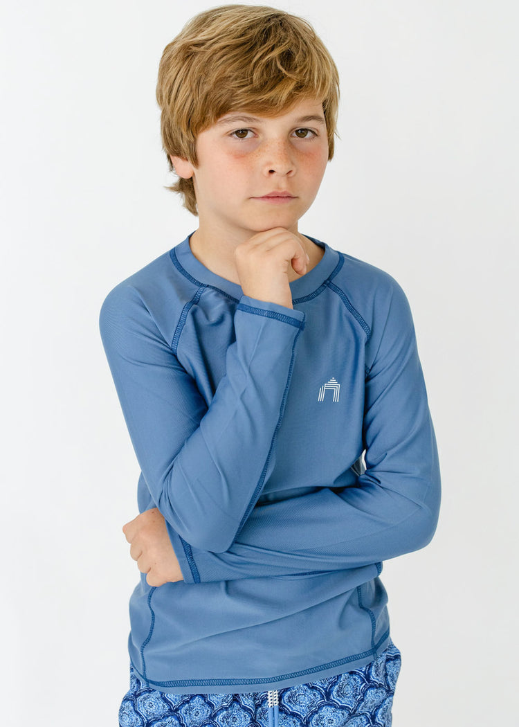 A blonde boy wearing the sun protective Boys Light Blue Long Sleeve Rashguard from Cabana Life.