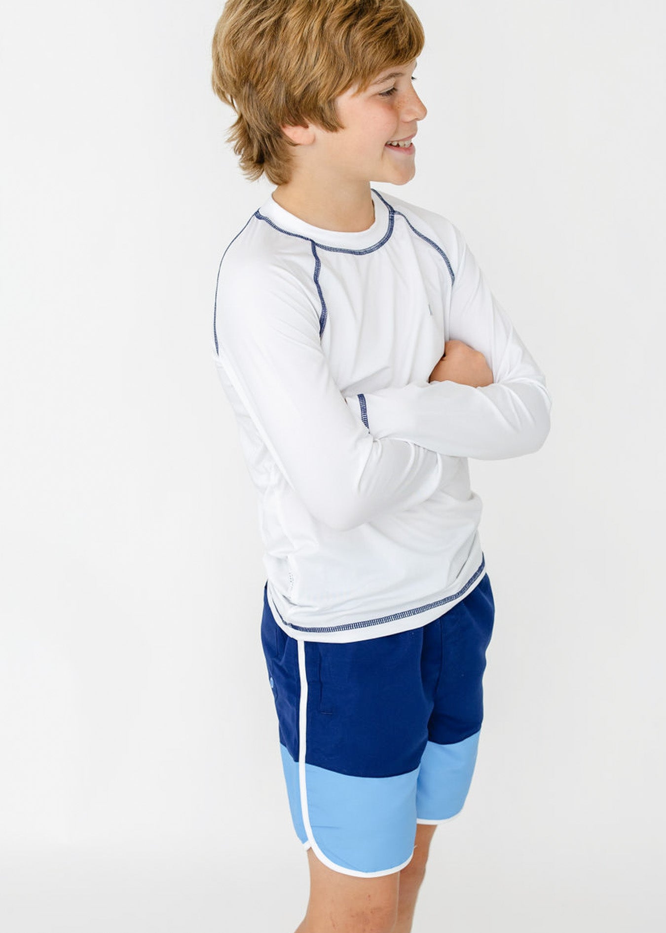 The side of a blonde boy with his arms crossed wearing the sun safe Boys White Long Sleeve Cabana Rashguard and Boys Blue Colorblock Swim Trunk from Cabana Life.