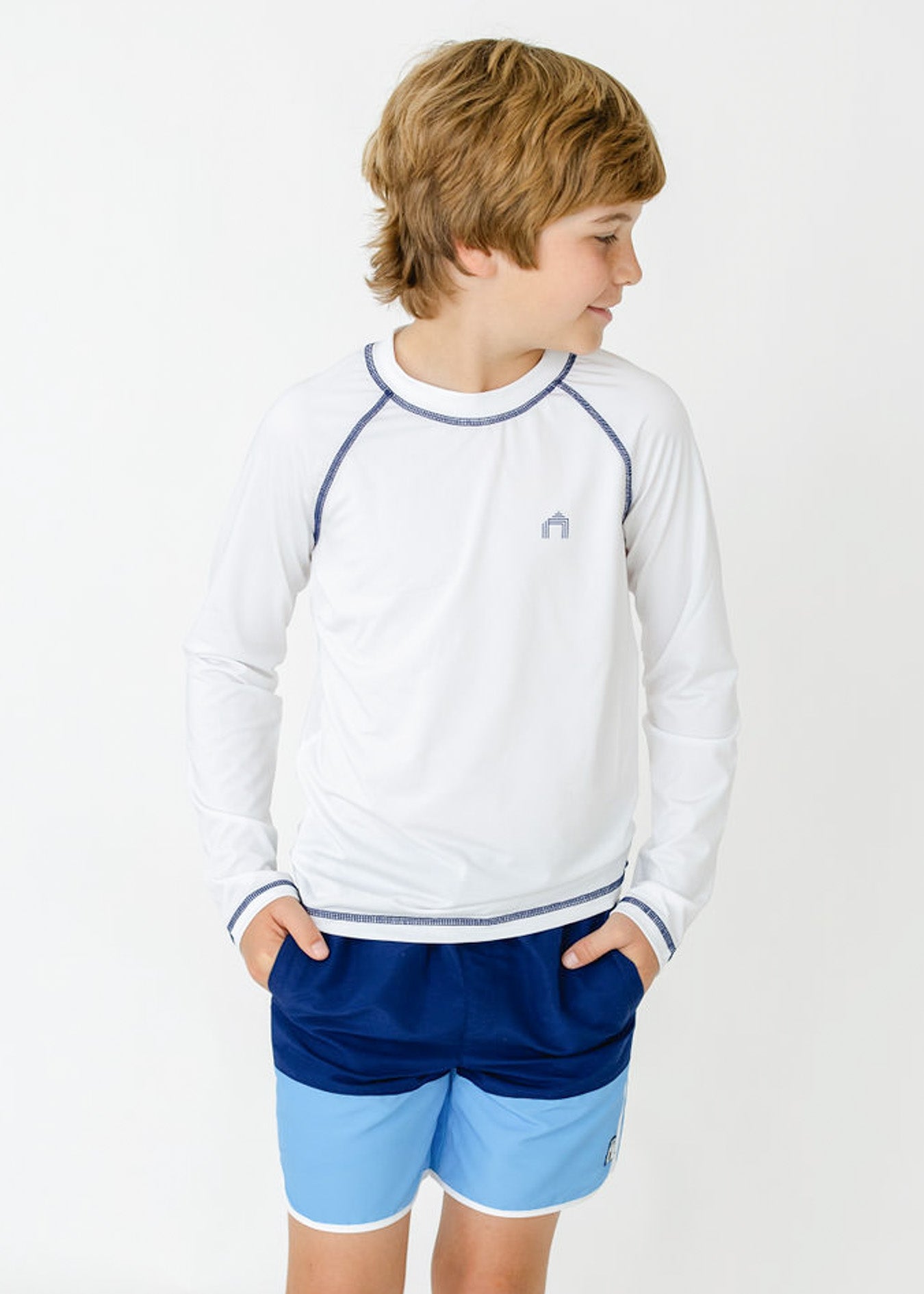 A blonde boy wearing the sun safe Boys White Long Sleeve Cabana Rashguard with hands in pockets of the Boys Blue Colorblock Swim Trunk from Cabana Life.