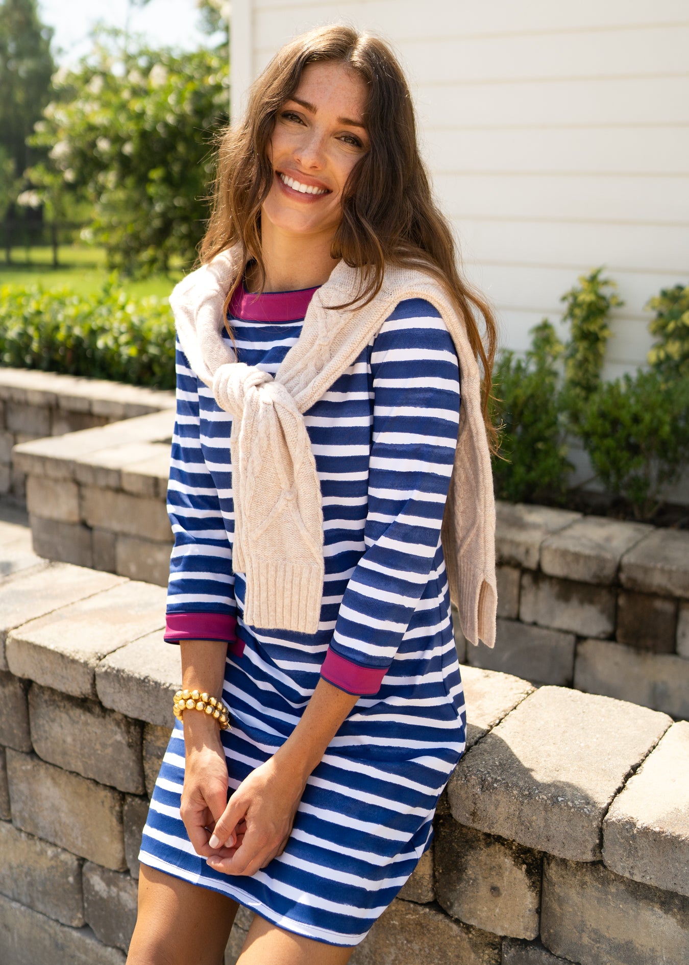 A brunette woman sitting on a brick wall wearing Cabana Life's sun protective Watercolor Stripe Cabana Shift Dress with a white sweater over her shoulders.