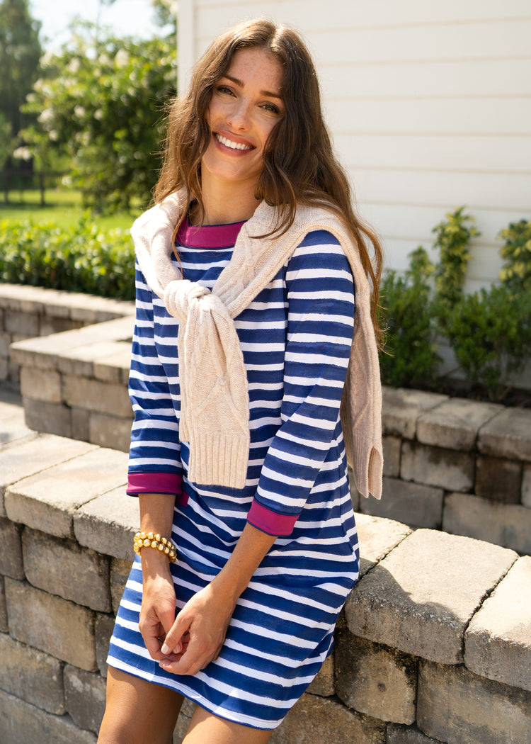 A brunette woman sitting on a brick wall wearing Cabana Life's sun protective Watercolor Stripe Cabana Shift Dress with a white sweater over her shoulders.