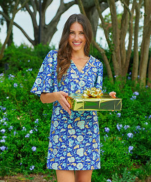 A brunette woman holding a gold present in front of a bush with blue flowers wearing the Cabana Life sun protective Navy Floral Metallic Embroidered Flutter Sleeve Shift Dress.