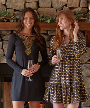 A brunette woman wearing the Cabana Life sun protective Black Metallic Keyhole Dress with a redhead woman wearing the Black Metallic Relaxed Dress in front of a fireplace holding champagne glasses.