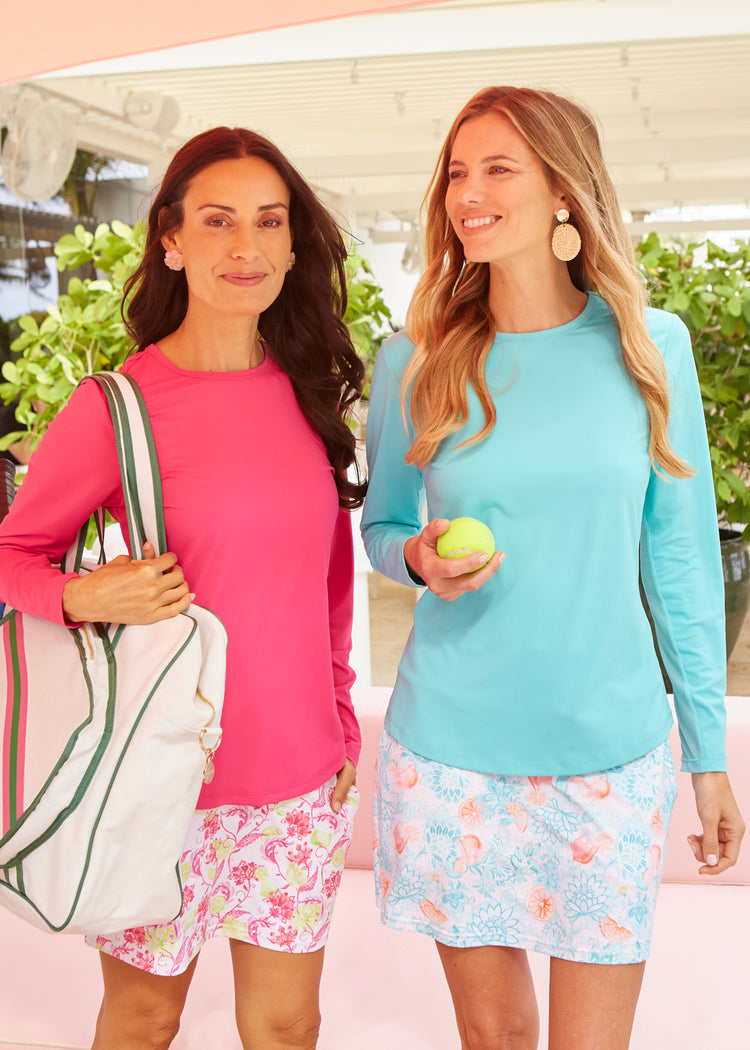 A brunette woman wearing a tennis bag, the Cabana Life sun protective Magenta Long Sleeve Performance Top and a Margarita 15" Skort with a blonde woman holding a tennis bag wearing the Aqua Long Sleeve Performance Top and the Spritz 15" Skort on a beach sand boardwalk.