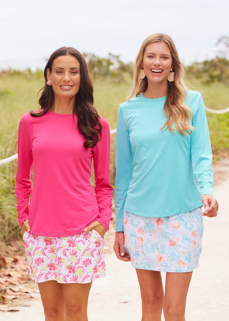 A brunette woman wearing the Cabana Life sun protective Magenta Long Sleeve Performance Top and a Margarita 15" Skort with a blonde woman wearing the Aqua Long Sleeve Performance Top and the Spritz 15" Skort on a beach sand boardwalk.