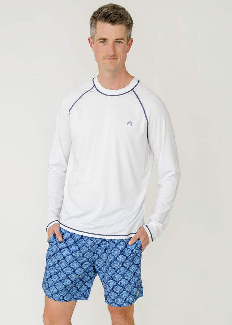 A man wearing the sun protective Men's White Long Sleeve Cabana Rashguard with hands in pockets of the Men's Seaside Swim Trunks from Cabana Life.