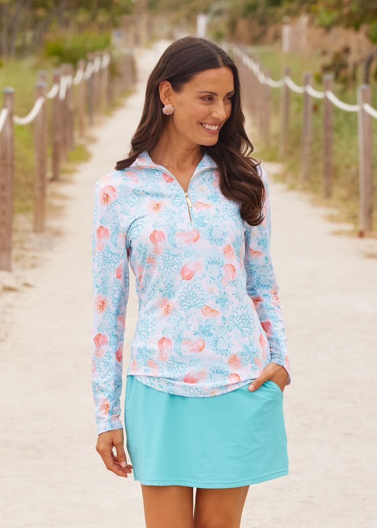 A brunette woman wearing Cabana Life's sun protective Spritz 1/4 Zip Sport Top with her hand in a pocket of the Aqua 15" Skort on a beach sand boardwalk.