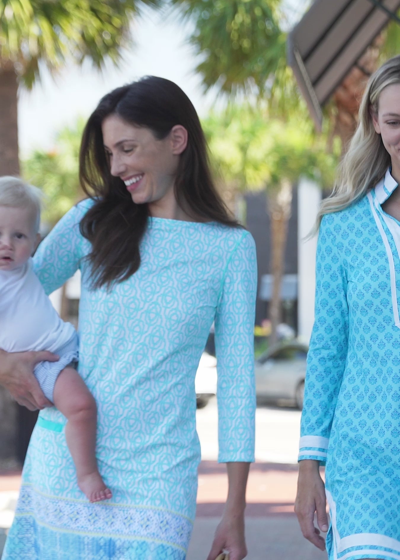 A brunette woman wearing the Cabana Life sun protective Coastal Cottage Cabana Shift Dress outside and holding a baby.